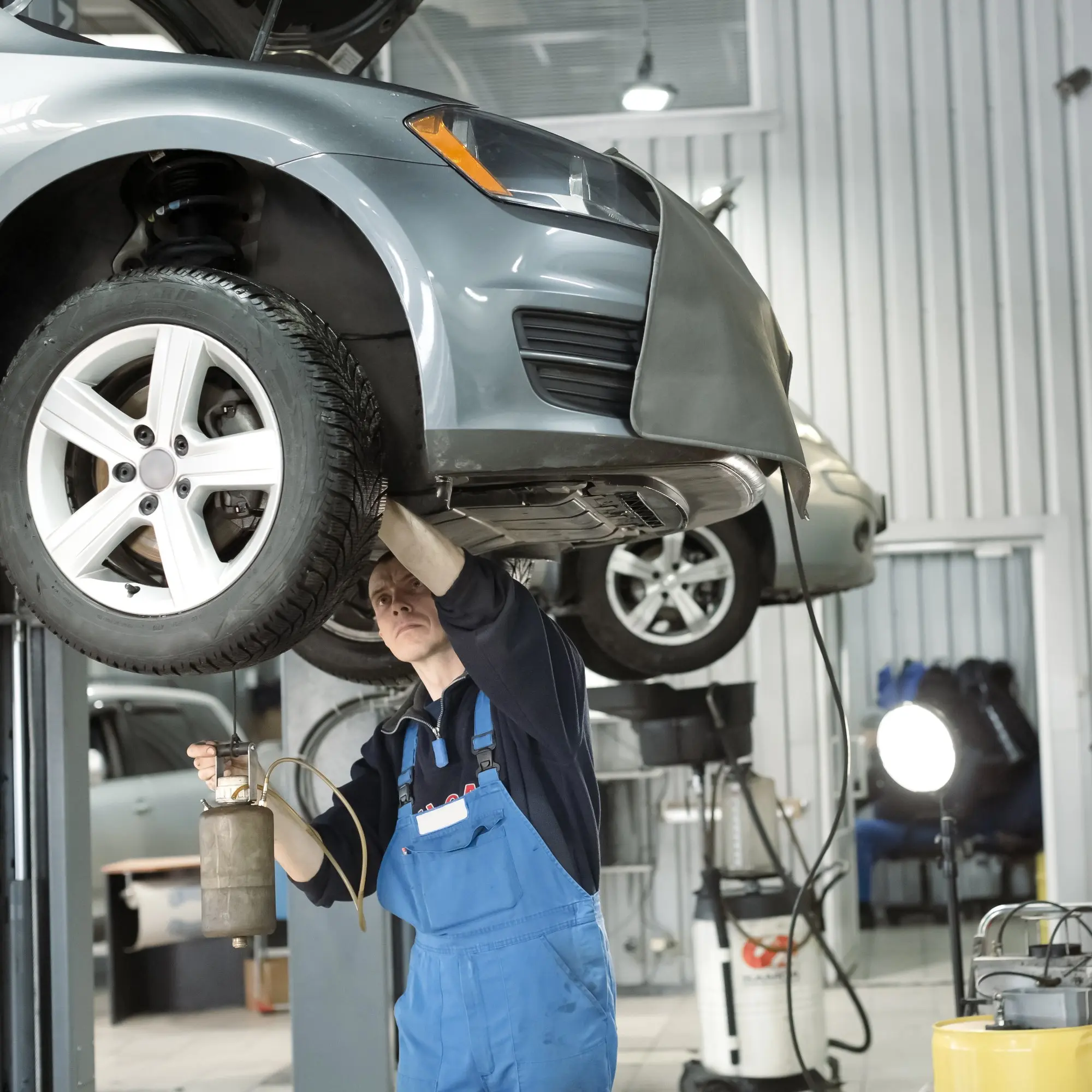 Mécanicien concentré en salopette bleue sous une voiture grise soulevée. Il effectue l'entretien à l'aide d'un outil de vidange dans un garage lumineux.