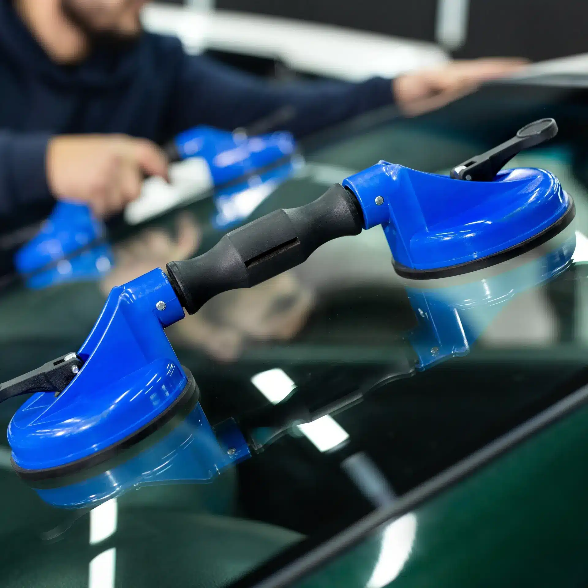 Outil double ventouse bleu fixé à un pare-brise de voiture. Un technicien effectue la pose du vitrage auto en arrière-plan.
