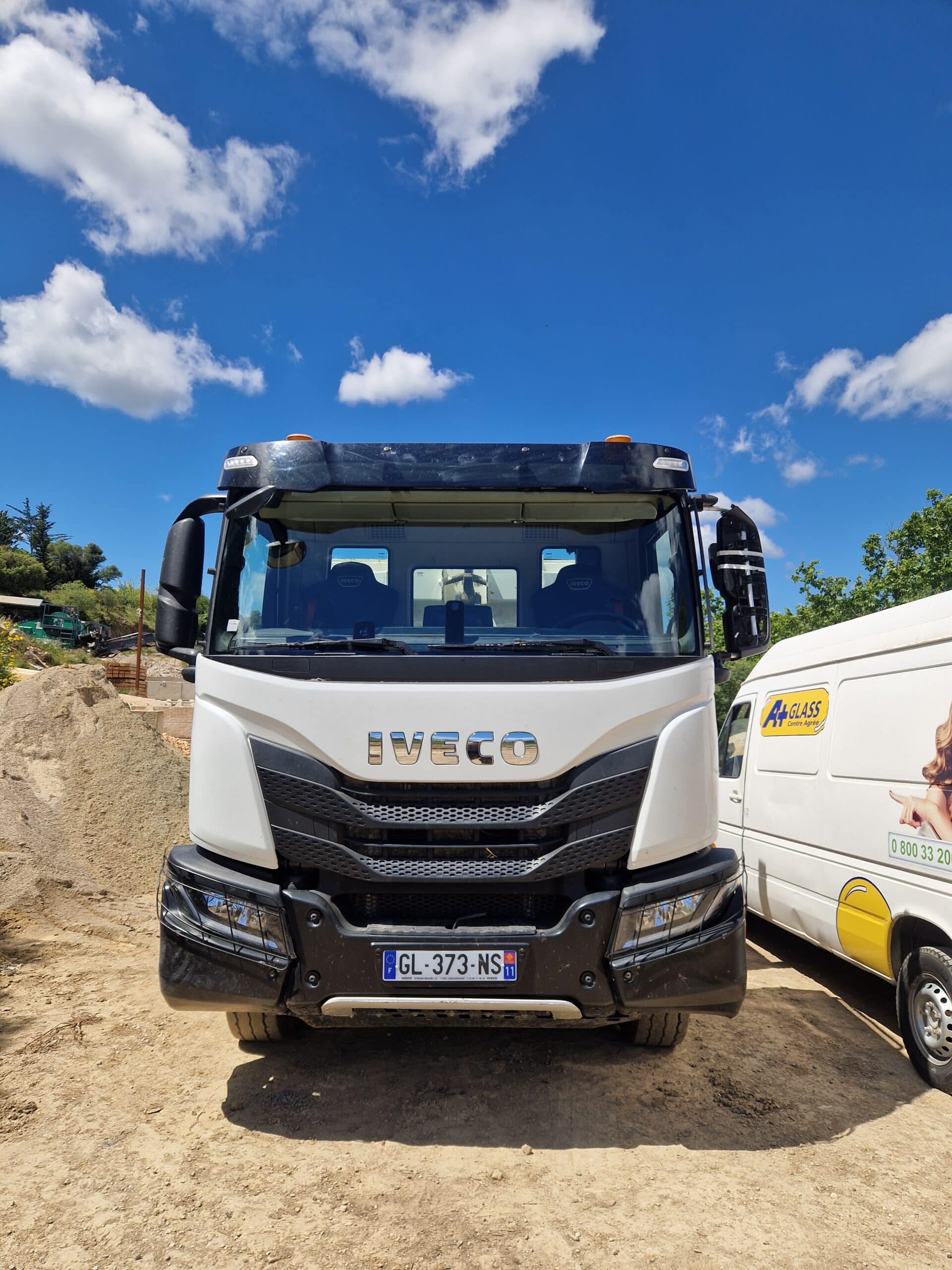 Camion Iveco blanc, plaque GL-373-NS, garé sur un chantier avec un fourgon A+ Glass sous un ciel bleu nuageux.