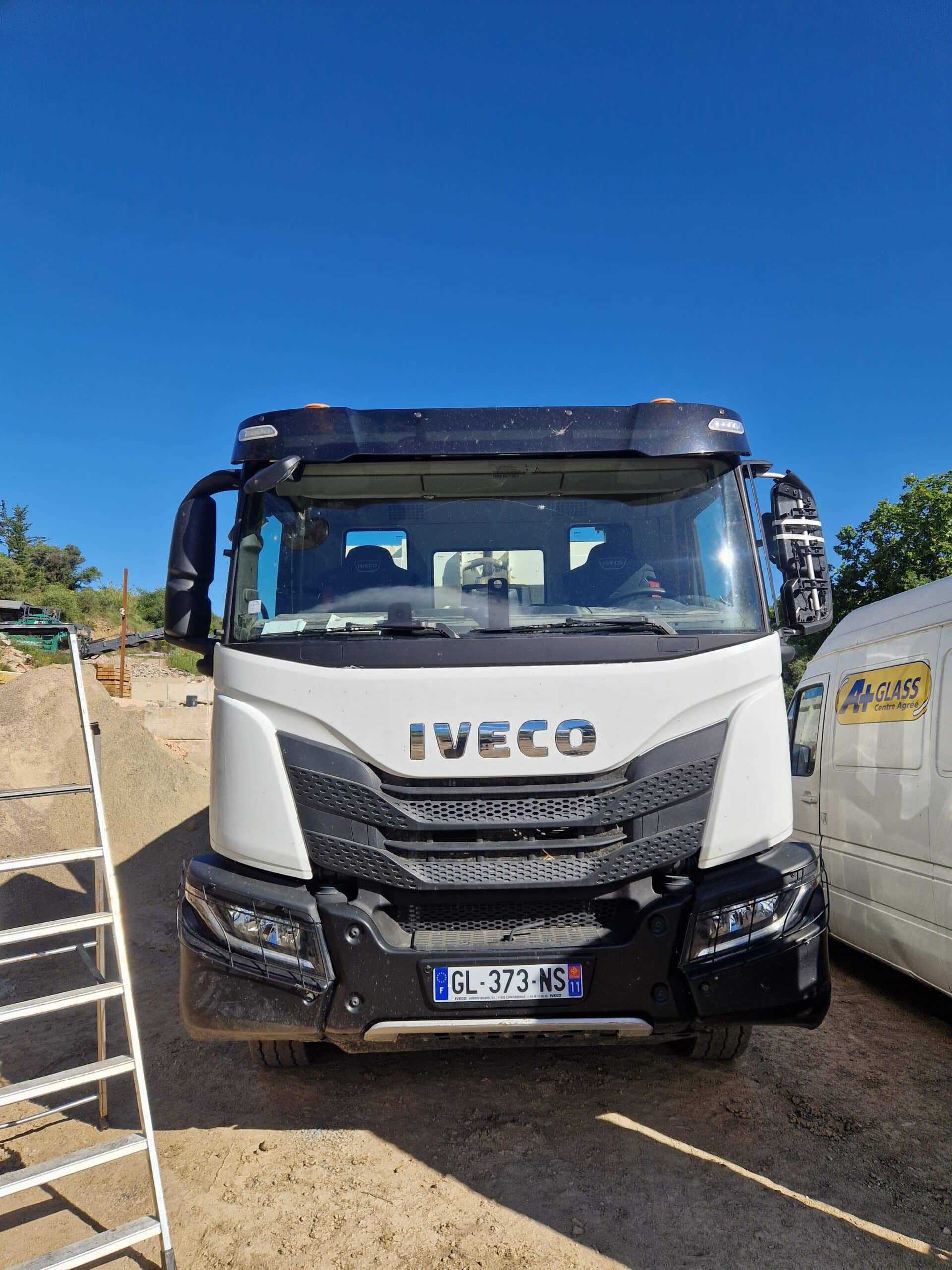 Camion Iveco blanc avec plaque GL-373-NS garé sur un chantier à côté d'un utilitaire A+ Glass sous un ciel bleu.