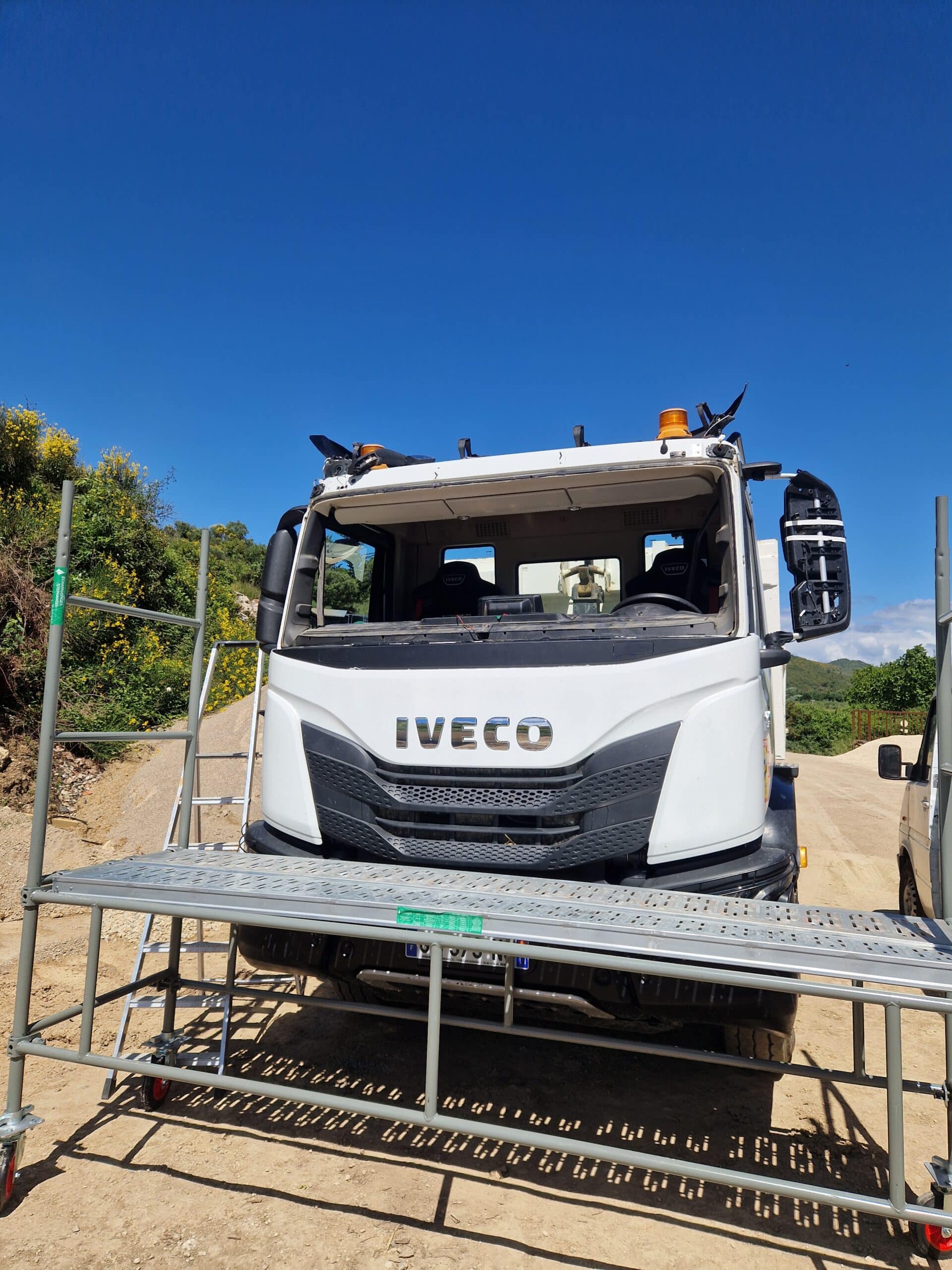 Camion Iveco blanc stationné derrière un échafaudage mobile sur un chantier de construction sous un ciel bleu.