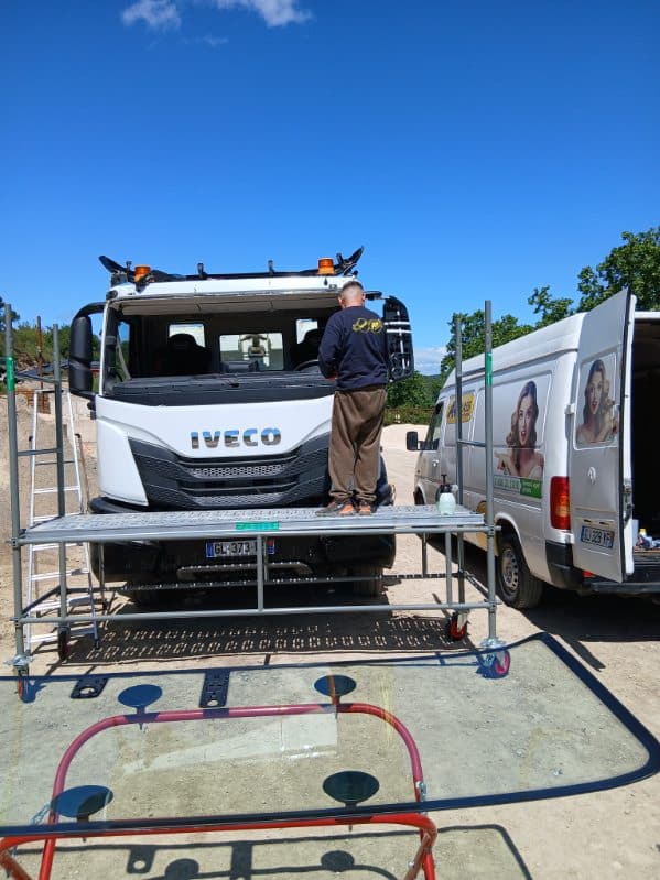 Technicien sur échafaudage installant un nouveau pare-brise sur un camion IVECO blanc lors d'une intervention.