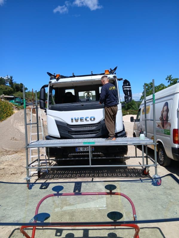 Technicien remplaçant un pare-brise de camion IVECO sur un échafaudage mobile à côté d'une camionnette Carglass.